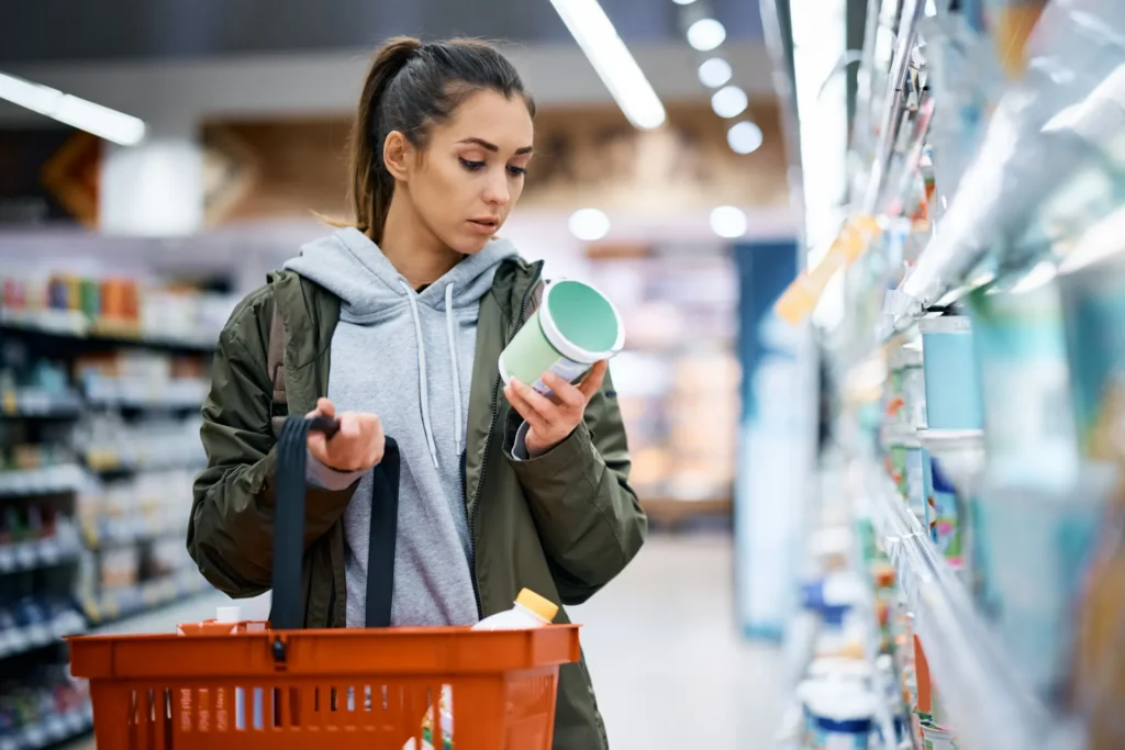 Ragazza al supermercato con un carrello della spesa su un braccio e con in mano un vasetto di yogurt.